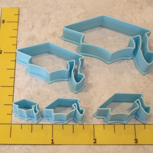 May include: Set of four blue plastic graduation cap cookie cutters. The cutters are in different sizes, with the largest cutter measuring approximately 2 inches in diameter.