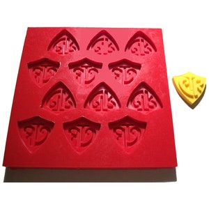 May include: A red silicone mold with 12 shield-shaped cavities, each featuring an ornate design. A separate yellow shield-shaped object is next to the mold. The mold and object are likely for crafting or baking.