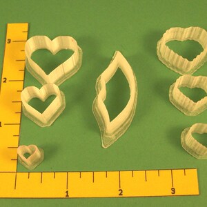 May include: A collection of clear plastic cookie cutters in various heart shapes and a leaf shape, arranged on a green surface. The cutters range in size, with the largest heart shape measuring approximately 3 inches across. A yellow ruler is in the image.