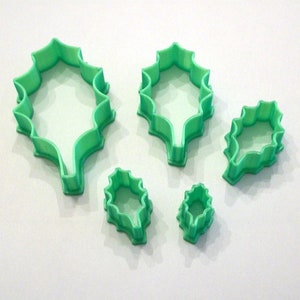 May include: Set of five green plastic holly leaf cookie cutters. The cutters are in various sizes, with the largest cutter being about 4 inches tall.