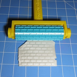 May include: A yellow and teal brick pattern roller with the text "LoveOfColor3D" is shown rolling over a piece of gray material, creating a brick pattern. The roller is on a blue grid surface.