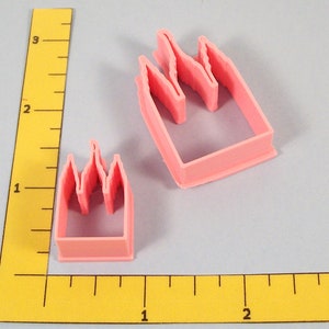 May include: Two pink plastic cookie cutters in the shape of a castle or cityscape. One is larger than the other. The cutters have a rectangular base with a crenelated top. The background is a light gray.
