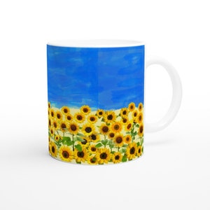 Puede incluir: Taza de cerámica blanca con un diseño de campo de girasoles bajo un cielo azul brillante. Los girasoles tienen pétalos amarillos y centros marrones. La taza tiene un asa blanca y un acabado brillante, ideal para bebidas calientes.