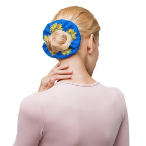 Puede incluir: Una goma para el pelo azul con un diseño de girasol amarillo. La goma está atada al cabello rubio de una mujer.
