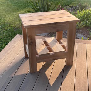 May include: A small, square wooden side table with a natural finish. The table has a slatted top and a lower shelf, supported by four sturdy legs. The table is approximately 20 inches tall and 18 inches wide, ideal for outdoor use.