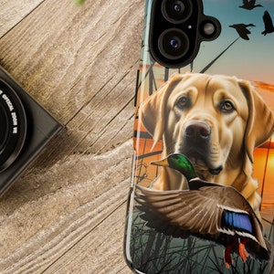 May include: A phone case with a yellow Labrador Retriever and a green duck. The background is a sunset with a lake and trees.