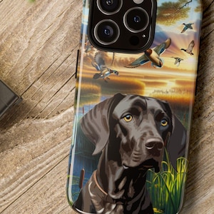 May include: A smartphone case featuring a black dog with yellow eyes, set against a backdrop of ducks in flight over a lake at sunset. The case has a glossy finish and is resting on a wooden surface.