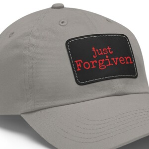 May include: A gray baseball cap with a curved brim. A black rectangular patch on the front displays the words "just Forgiven" in red, with a white border. The cap is made of a soft fabric.