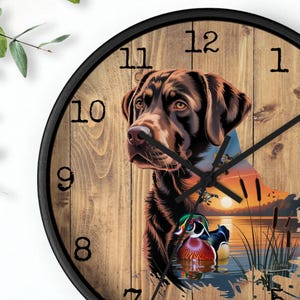 May include: Round wall clock with a black frame. The clock face features a brown Labrador Retriever, a sunset scene, and a colorful duck. The background has a wood-grain texture with black hands and numerals.