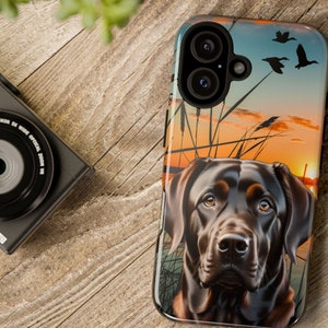 May include: A black Labrador Retriever dog with brown eyes is featured on a phone case. The dog is looking directly at the camera. The background is a sunset scene with ducks flying over a lake.
