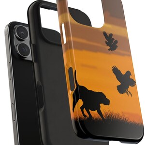 May include: A phone case featuring a sunset scene with a black dog silhouette and flying birds. The case has a black interior and a colorful exterior with a dog and birds in silhouette.