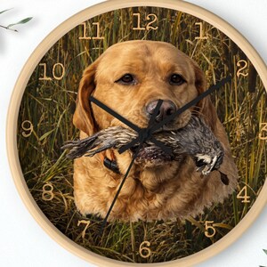 May include: A round wall clock with a light wood frame. The clock face features a golden retriever holding a bird in its mouth, set against a backdrop of tall grass. Black clock hands and numerals complete the design.