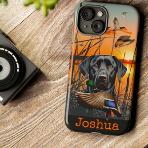 May include: Black phone case featuring a black Labrador Retriever holding a duck, set against a sunset scene with flying ducks and reeds. The name "Joshua" is printed in orange at the bottom. The case is on a wooden surface.