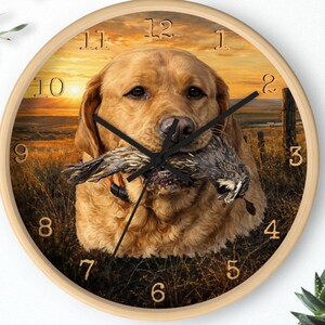 May include: A round wall clock with a light wood frame. The clock face features a golden retriever holding a bird in its mouth, set against a sunset backdrop. Black clock hands and gold numerals complete the design.