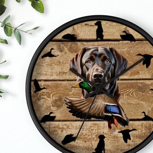May include: A black Labrador Retriever with a duck in its mouth, on a wooden background with silhouettes of ducks flying around the dog. The clock face is visible with black hands.