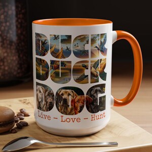 May include: White ceramic mug with an orange rim and handle. The mug features a colorful illustration of ducks and dogs with the words "Duck Duck Dog" and "Live - Love - Hunt" printed on the front.