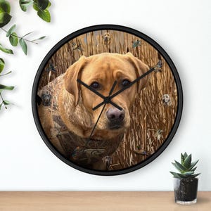 May include: Round wall clock with a black frame. The clock face features a yellow Labrador Retriever with a camouflage pattern and ducks. The clock hands are black.