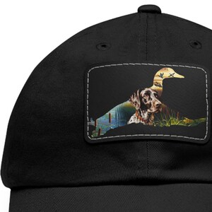 May include: A black baseball cap with a patch featuring a brown and white dog silhouette with a duck flying overhead. The patch has a green, brown, and blue background with reeds and water.