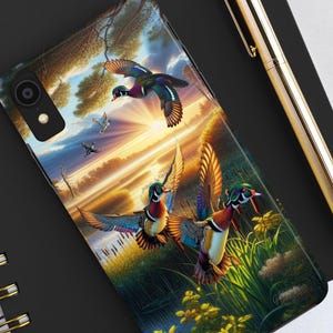 May include: A phone case with a colorful image of three wood ducks flying over a lake at sunset. The ducks are in flight with their wings spread. The background is a vibrant sunset with a blue sky and orange clouds. The lake is surrounded by green reeds and trees.