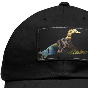 May include: A black baseball cap with a patch featuring a brown dog silhouette with a duck silhouette superimposed over it. The dog is lying down with its head turned to the side. The duck is flying with its wings spread. The patch has a white outline and is set against a black background.