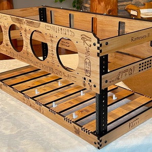 May include: A two-tiered wooden shelf with a light brown finish and black accents. The shelf features circular cutouts, engraved hieroglyphs, and the text "Rameses II". The structure is supported by black metal posts and includes white pegs.