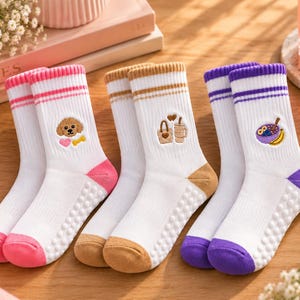 Könnte beinhalten: Drei Paar weiße Socken mit gerippten Bündchen und farbigen Streifen. Jedes Paar zeigt ein kleines gesticktes Motiv: einen braunen Hund mit Herz und Knochen, eine beige Einkaufstasche mit Getränk oder eine violette Schale mit Obst. Fersen und Zehen sind rosa, beige oder violett, die Sohlen haben weiße Noppen.