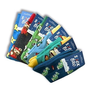 Peut inclure: Une collection de chaussettes fantaisie avec des motifs sur le thème des jeux vidéo. Les chaussettes sont de couleurs bleu, vert, gris et rouge, avec des personnages et des motifs. Chaque paire a une étiquette bleue avec "SOCK BRO Novelty Sock Collection".