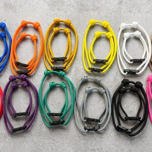 May include: A collection of colorful adjustable bracelets in various shades, including blue, orange, yellow, white, tan, red, purple, green, gray, black, and pink. Each bracelet features a black plastic clasp and knotted design.