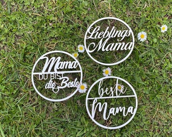 May include: Three white wooden circles with the words "Mama du bist die Beste", "Lieblings Mama", and "Beste Mama" written on them. The circles are on a bed of green grass with white flowers.