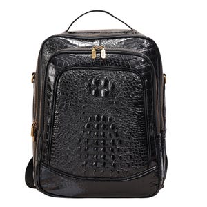 Puede incluir: Mochila negra con textura de piel de cocodrilo. La bolsa tiene un asa superior, un bolsillo frontal y cremalleras doradas. La mochila tiene forma rectangular y un acabado brillante. La bolsa tiene la espalda acolchada y correas para los hombros.