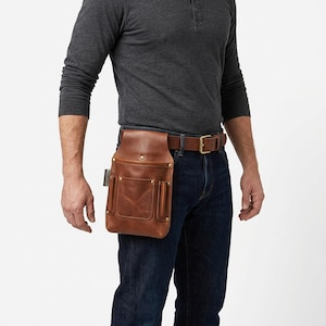 May include: A brown leather tool pouch featuring a large main compartment and two smaller front pockets, secured with brass-colored rivets. The pouch is attached to a brown leather belt with a metal buckle, worn around the waist over dark blue jeans.