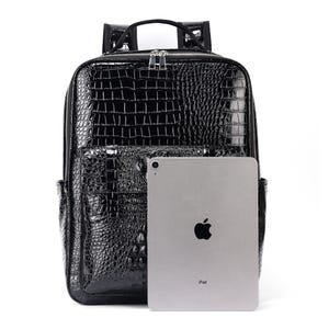 Puede incluir: Mochila negra con textura brillante de piel de cocodrilo. La mochila tiene un asa superior, un bolsillo frontal y una cremallera plateada. Un iPad se coloca junto a la mochila para comparar el tamaño.