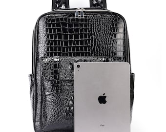 Pode incluir: Mochila preta com textura brilhante de pele de crocodilo. A mochila tem uma alça superior, um bolso frontal e um zíper prateado. Um iPad é colocado ao lado da mochila para comparação de tamanho.