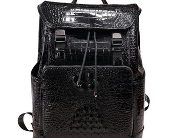 Pode incluir: Uma mochila preta com textura brilhante de pele de crocodilo. A bolsa apresenta um fecho de aba com fivelas de metal, um cordão e uma alça superior. Possui um bolso frontal e um bolso lateral.