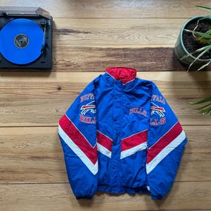 Puede incluir: Chaqueta azul, roja y blanca de los Buffalo Bills con logotipos y letras del equipo. La chaqueta tiene cuello y detalles en rojo, con rayas blancas. Un tocadiscos vintage y una planta en maceta están en el fondo.