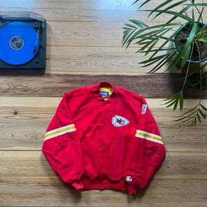 Puede incluir: Una chaqueta bomber roja de los Kansas City Chiefs con un patrón de rayas blancas y amarillas en las mangas, un logotipo de punta de flecha blanca en el pecho, un pequeño logotipo en la manga izquierda, forro interior amarillo y cierre frontal.