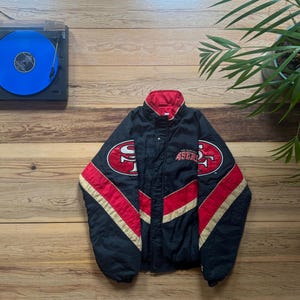 May include: A black, red, and gold San Francisco 49ers jacket featuring a high collar, a front snap-button closure, and embroidered team logos on the chest. The jacket has long sleeves with diagonal red and gold stripes and a red inner lining.