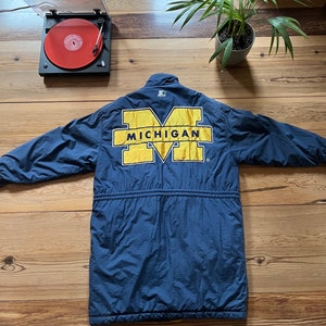 May include: A navy blue long-sleeved jacket with a large yellow "M" on the back. The "M" is outlined in white and has the word "MICHIGAN" written across the top in yellow.
