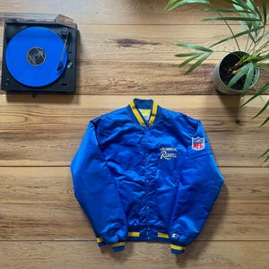Puede incluir: Chaqueta de satén azul de Los Angeles Rams con ribete amarillo, un logotipo de Starter y un parche de la NFL. Se ve un tocadiscos vintage con un disco de vinilo azul. La chaqueta está sobre una superficie de madera.