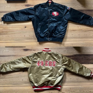Könnte beinhalten: Eine wendbare Satin-Bomberjacke mit einer schwarzen Seite, die ein San Francisco 49ers-Logo auf der Brust und einen NFL-Aufnäher am Ärmel zeigt, sowie einer goldenen Seite mit dem in Rot und Weiß gestickten Schriftzug 49 ERS auf dem Rücken.