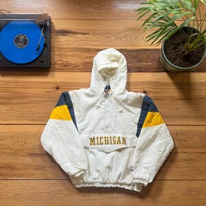 Puede incluir: Chaqueta blanca con capucha y detalles en amarillo y azul marino. La chaqueta lleva la palabra "MICHIGAN" bordada en el pecho. En el fondo, un tocadiscos con un disco de vinilo azul.