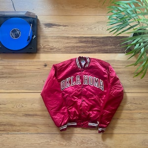 May include: A red satin jacket with the text "OKLA HOMA" in white block letters. The jacket has white and black striped trim on the collar, cuffs, and hem. A blue vinyl record is on a turntable.