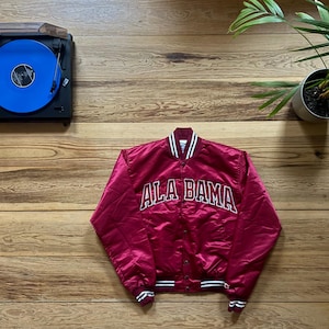 May include: A maroon satin varsity jacket with the word "ALA BAMA" in white lettering. The jacket has white and black striped trim on the collar, cuffs, and hem. A black record player with a blue record is in the background.