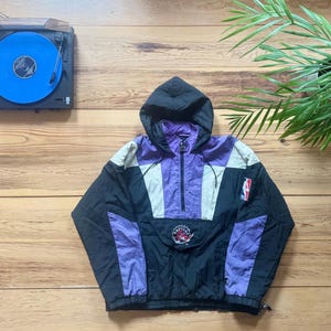 May include: A black, purple and white windbreaker jacket with a zip fastening and a hood. The jacket features a team logo on the front and a basketball logo on the sleeve.