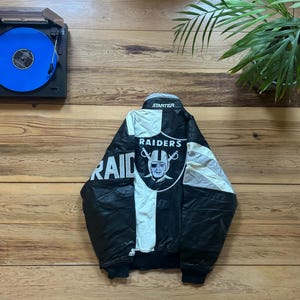 Peut inclure: Veste noire et blanche Raiders Starter avec le logo de l'équipe sur le devant et le mot "RAIDERS" sur la manche. La veste est sur une surface en bois avec un tourne-disque et un disque bleu.