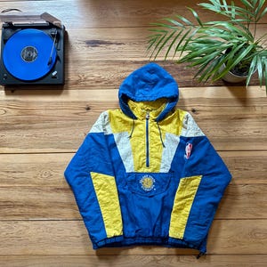 Könnte beinhalten: Eine blaue, gelbe und weiße Windbreaker-Jacke mit einem Golden State Warriors-Logo auf der Vorderseite. Die Jacke hat einen Reißverschluss und eine Kapuze.