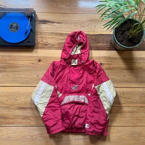 May include: A vintage red and cream-colored Starter anorak jacket with a hood. The jacket features a front pocket with the word "Minnesota" embroidered on it. A record player with a blue vinyl record is in the background.