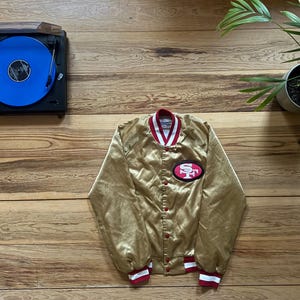 Könnte beinhalten: Goldene Satinjacke mit roten und weißen Zierleisten, mit einem San Francisco 49ers Logo-Patch. Die Jacke ist zugeknöpft und liegt auf einer Holzoberfläche. Im Hintergrund befindet sich ein Plattenspieler mit einer blauen Schallplatte.