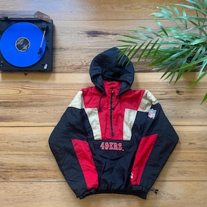 Könnte beinhalten: Eine schwarze, rote und weiße Windbreaker-Jacke mit Reißverschluss und Kapuze. Die Jacke trägt den Schriftzug "49ERS" auf der Vorderseite und ein NFL-Logo auf dem Ärmel.