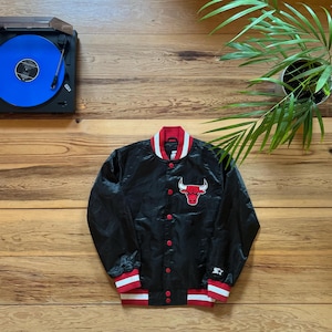 Può includere: Giacca bomber nera in satin con costine a righe rosse e bianche al collo, ai polsini e all'orlo. La giacca presenta un logo ricamato rosso dei Chicago Bulls sul petto sinistro.